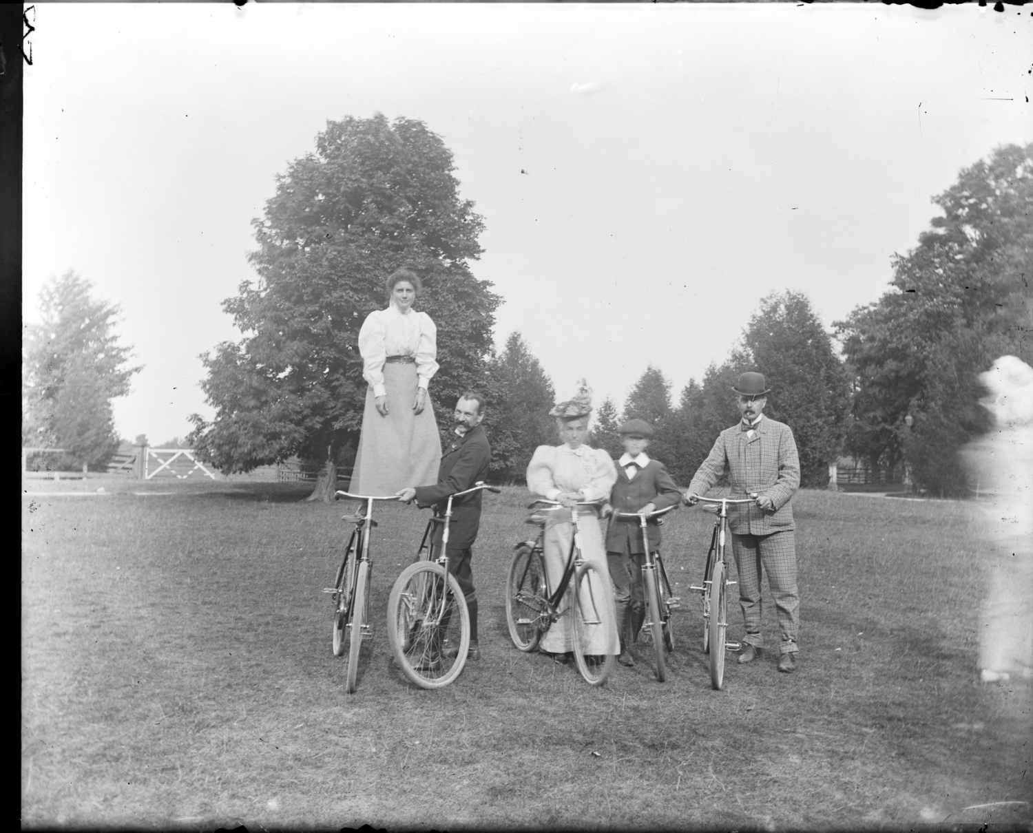 A photograph of a group of people posing with bicycles on the property of the Patteson family in Eastwood, Ontario. One young woman in the photo is standing on the seats of two bicycles.