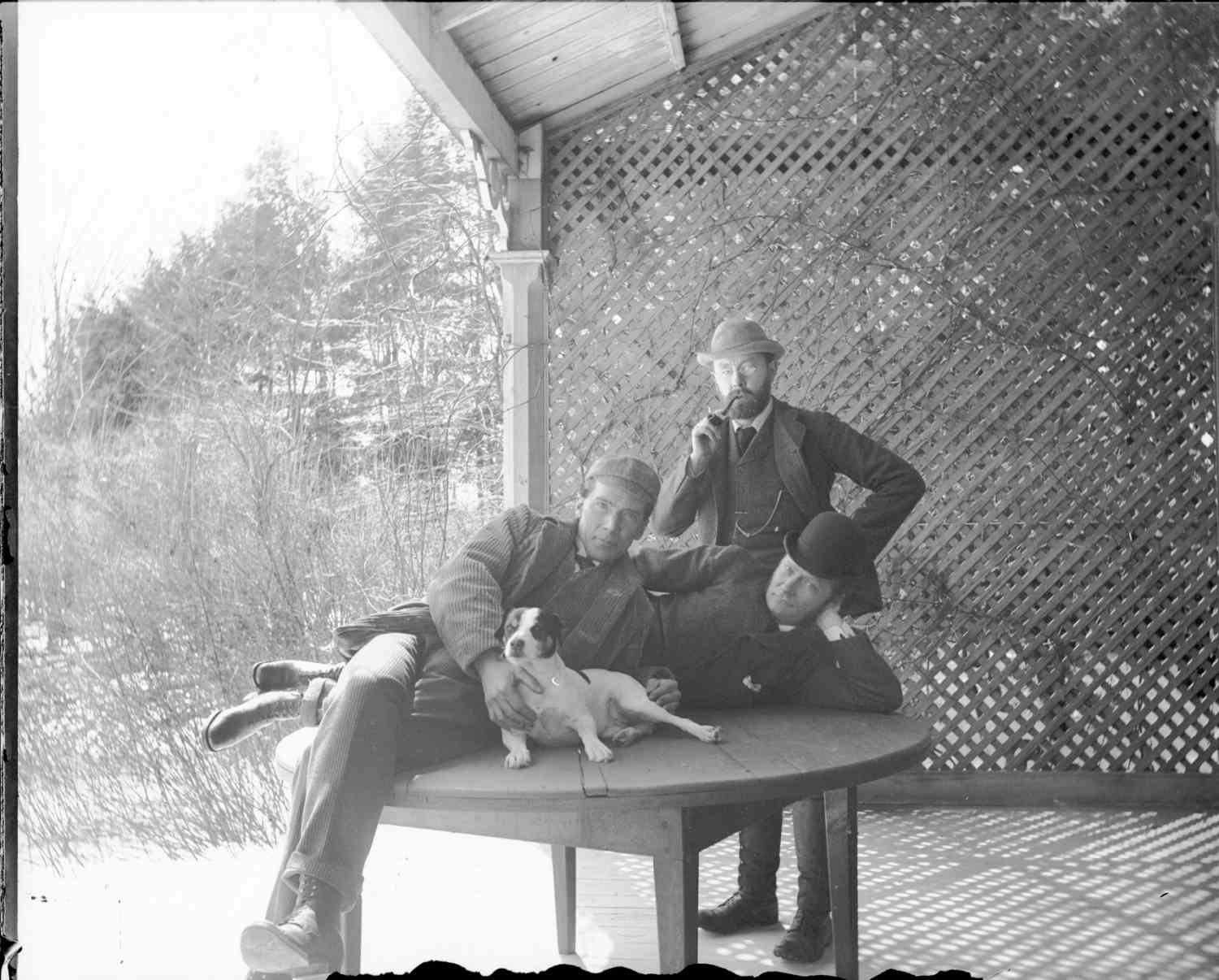 Two men are laying on a wooden table outside in a sheltered area. The men are wearing suits and hats and have a small dog laying on the table in front of them. A man is standing behind the table, also wearing a suit and bowler hat, he is wearing glasses, has a beard, and is smoking a pipe.