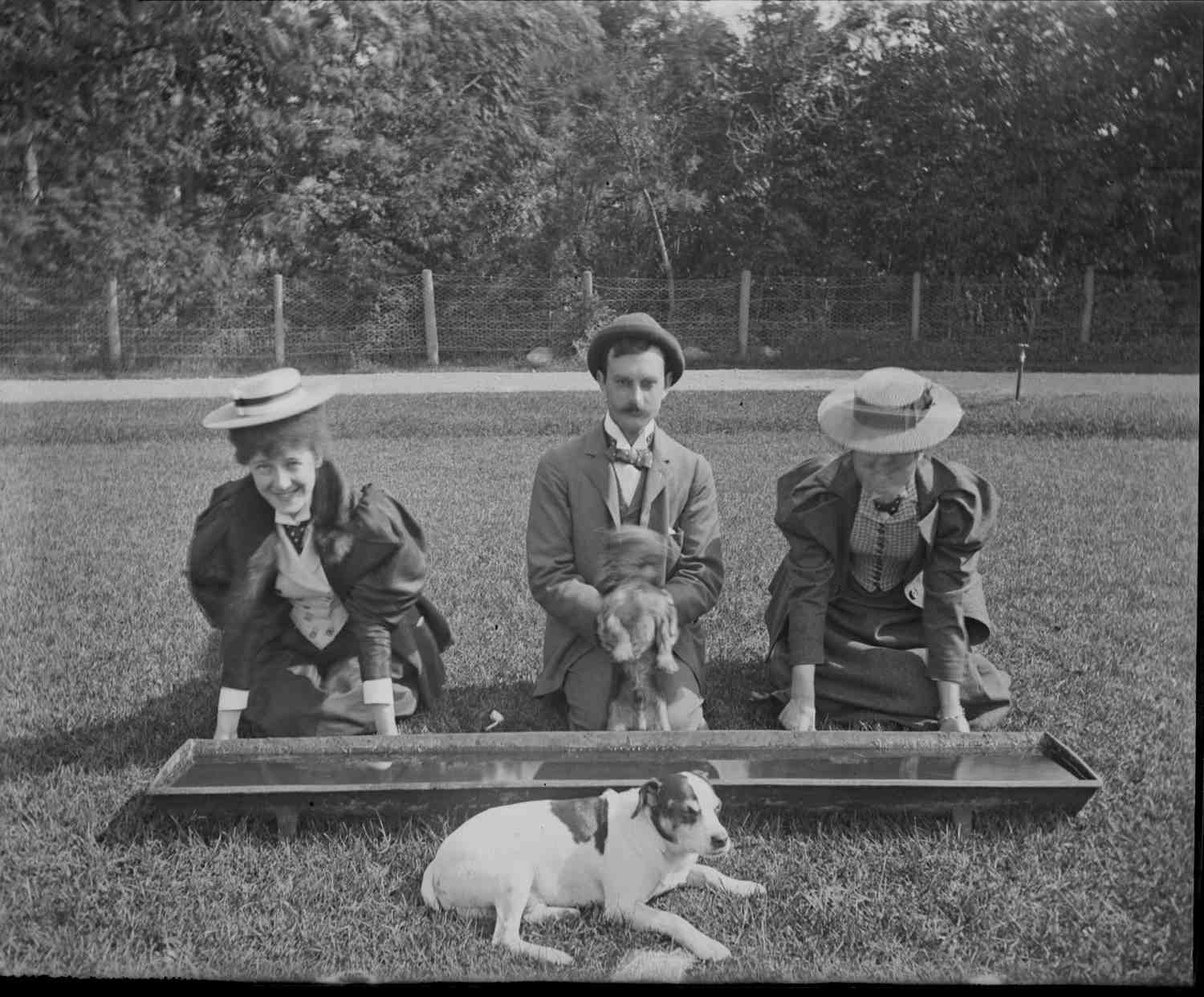 A man and two women pose kneeling in front of a water trough, pretending to drink from it like livestock.