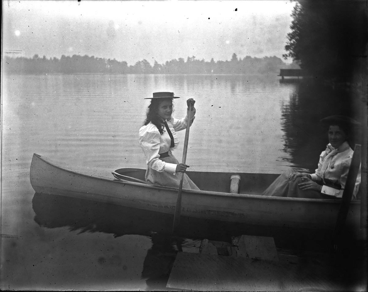 Two women wearing dresses and hats sit in canoes on a lake.