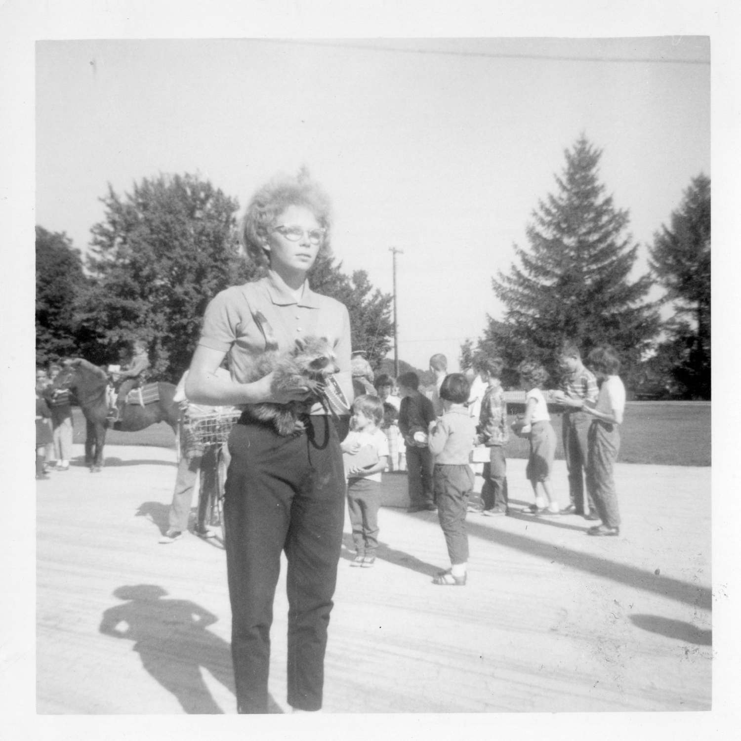 Person standing in the middle of a pet show and holding a small raccoon. The person is wearing a polo shirt with trousers, and wearing eye glasses. There are many children in the background, one child is riding a pony.