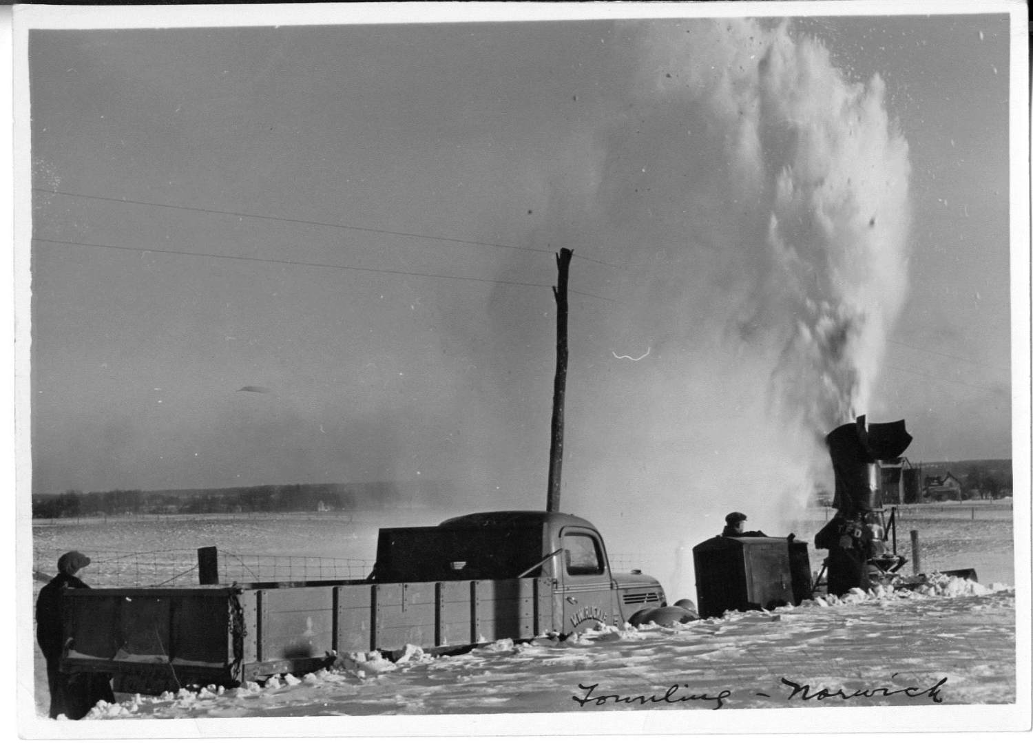 Two men use a plow to clear deep snow from a road.