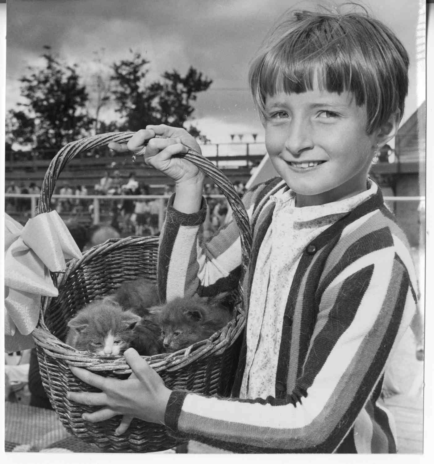 A young girl holding a basket of small kittens at a pet show. The girl is smiling. People are gathered on bleacher seats behind her.