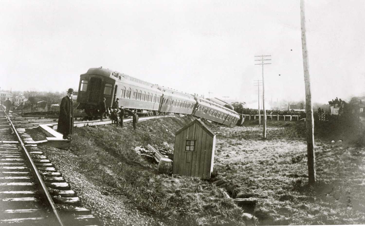 A photo of the Lehigh Express train derailment. The train car are off the rails. A group of people are standing at the back of the train. One man is in the foreground wearing a long coat and bowler hat.