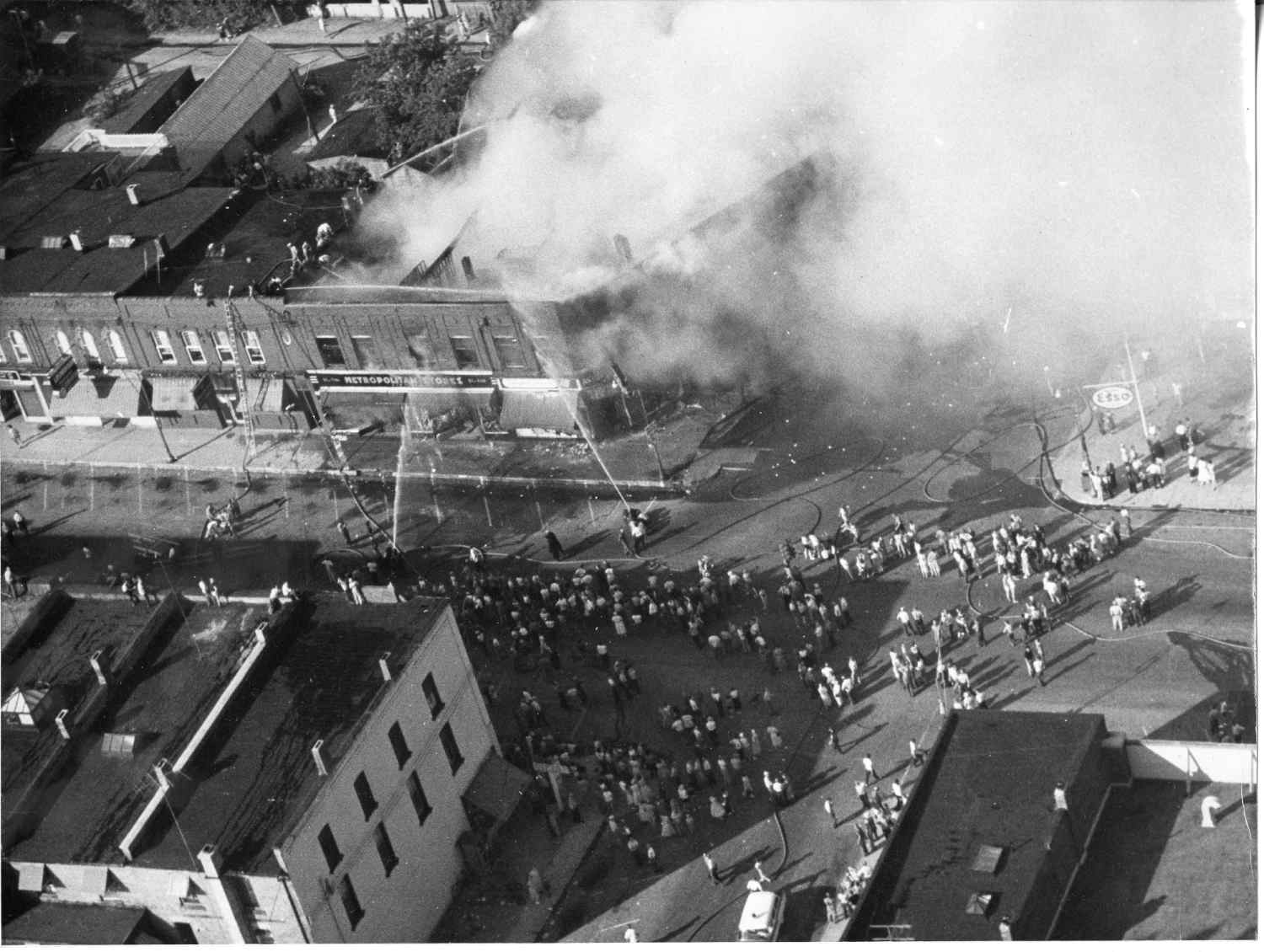 Aerial view of downtown Tillsonburg, black and white photo. A large crowd is gathered in the middle of an intersection. Firefighter stand on the street using hoses to put out a fire. A building is on fire, a large amount of smoke is billowing out of the building.