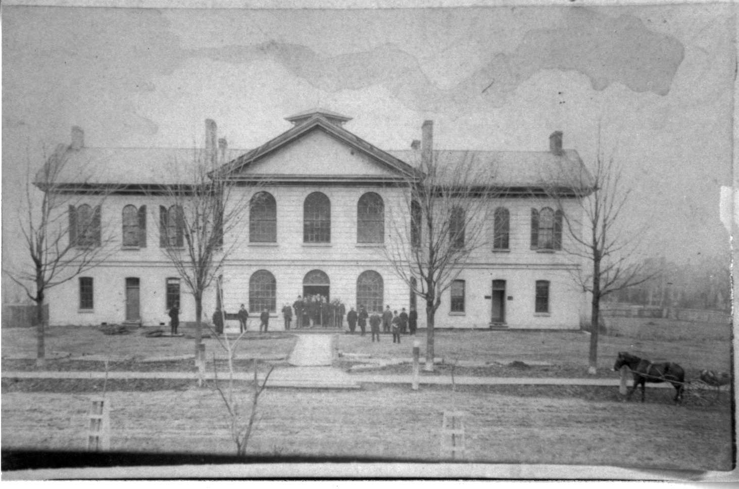 A black and white photograph of the original county courthouse building in Woodstock, Ontario. A group of men are standing outside of the building. It is a very large two-story building with arched windows and four leafless trees on the front lawn.