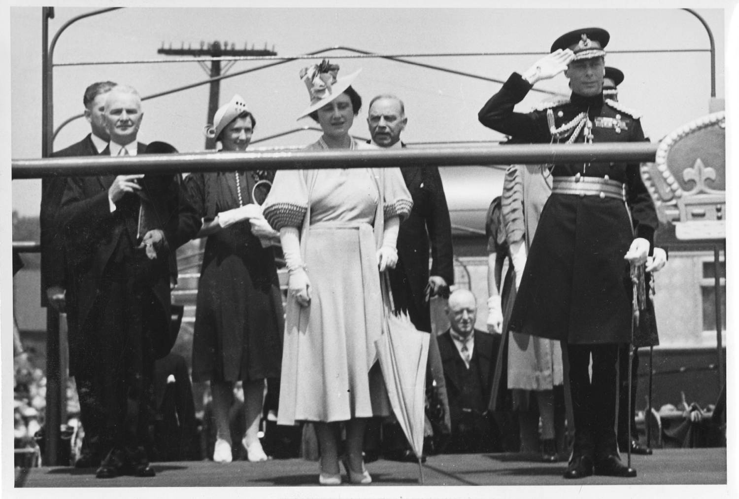 King George VI and Queen Elizabeth (the Queen Mother) stand on a platform at the CNR station in Woodstock. The queen is wearing a light coloured dress and a hat with flowers in it. The king is wearing a military uniform and is saluting. A few other people stand behind them.