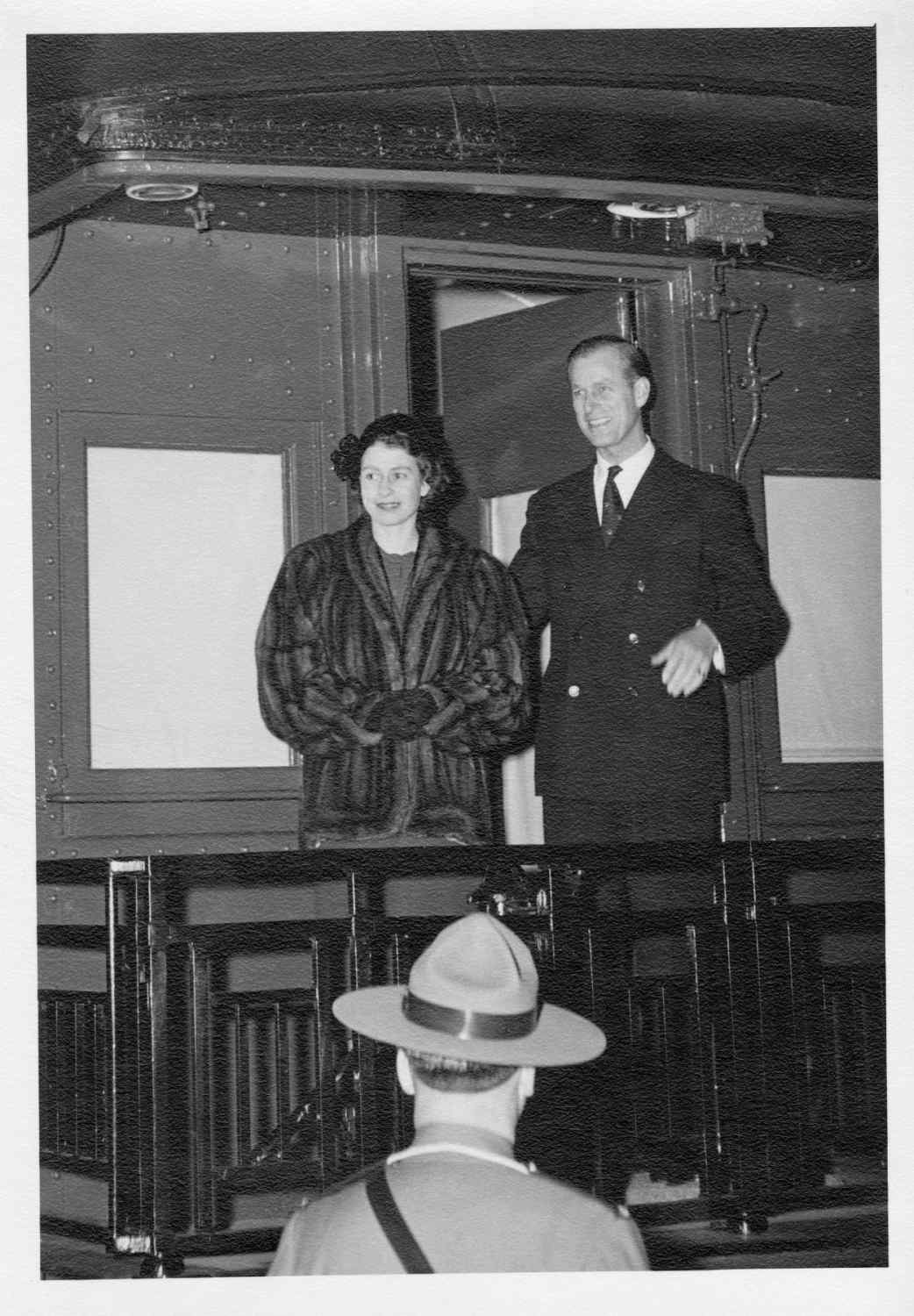 Princess Elizabeth and Prince Philip, the Duke of Edinburgh, stand on the back of a train. Princess Elizabeth was wearing a fur coat and Prince Philip was dressed in a suit.