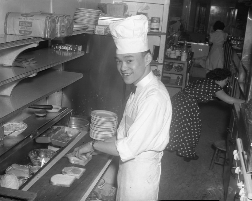 Hugh Chong, an Asian-Canadian man, wearing a work uniform and chef's hat, working in the kitchen of his family owned Food Rite Restaurant in Woodstock, Ontario. Mr. Chong appears to be making sandwiches. Two East Asian women are working in the background of the photo.