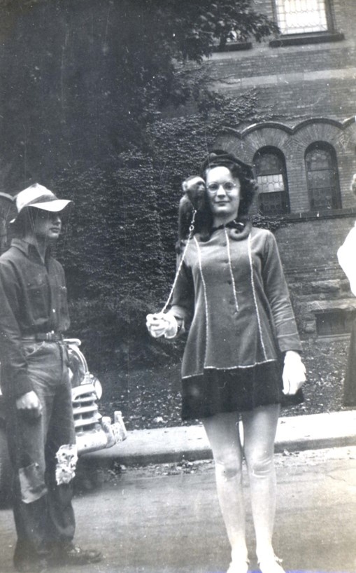 A young woman named Barbara Brown is posing with her pet monkey on her shoulder. She is wearing a dress with stocking, white shoes, and white gloves. Standing to the left is a man wearing a floppy hat, a shirt, and pants with patches on the knees. At Woodstock Collegiate Institute, for field day.