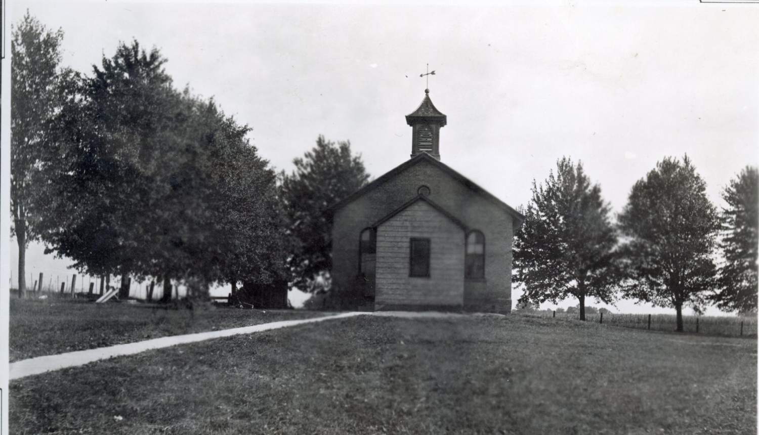 A black and white photograph of the old public school in Hickson, Ontario, Canada. It is a brick, one-room schoolhouse. A bell and weather vane sit on top of the school roof.