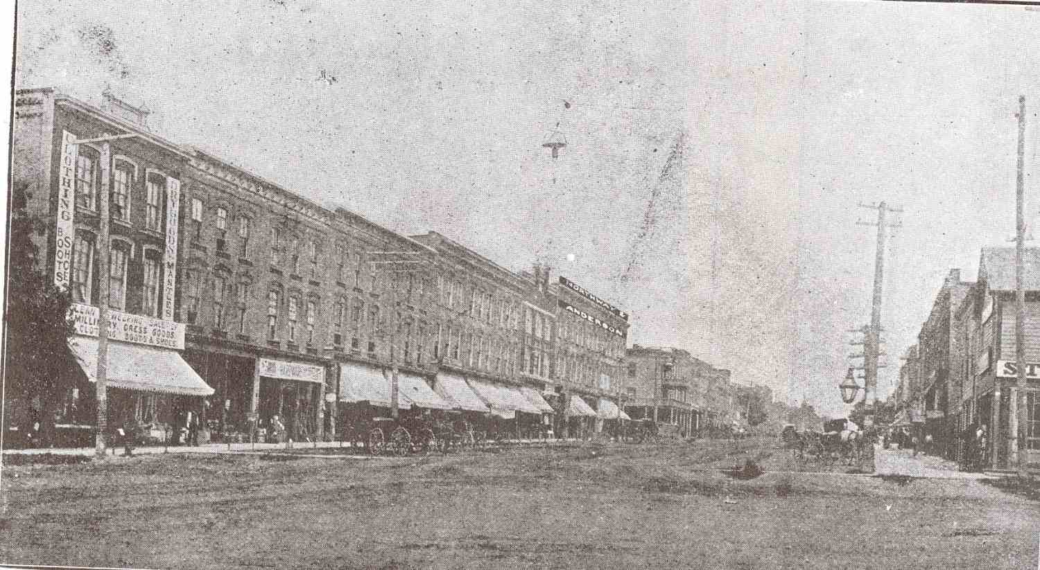 A street view of Broadway Street in Tillsonburg. West side of the street looking north. Brick and wood frame buildings line the dirt road, shop and business signs mounted on the front of the buildings. Hydro poles, carriages, and horse drawn wagons also line the street.