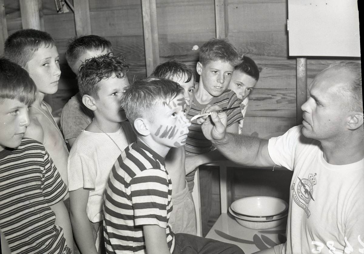 A man paints a boy's face with a group of other boys surrounding them and watching.