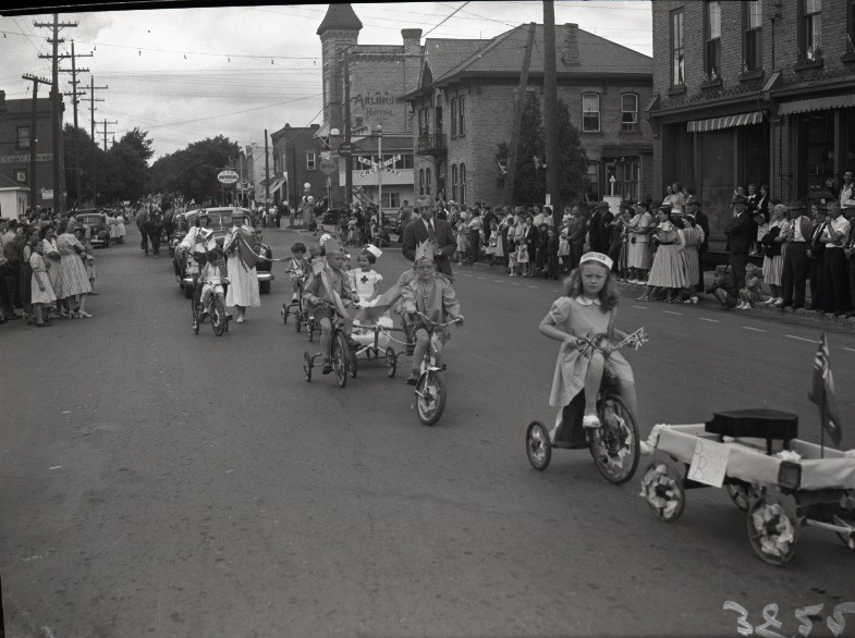 Children wearing costumes and riding tricycles in a parade in Tavistock, Ontario. Cars follow behind them and crowds of people are lined up along the sides of the street to watch.