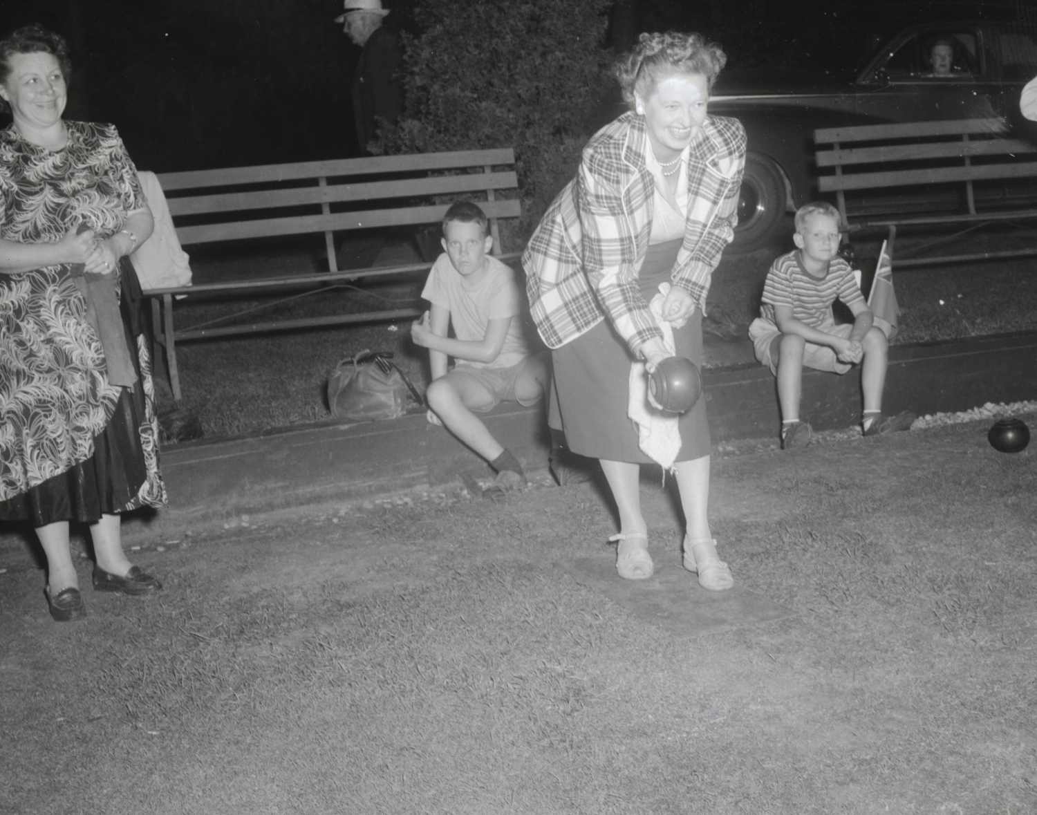 A woman lawn bowling with a smile on her face. A woman is standing off to the side watching her along with two boys in the background.