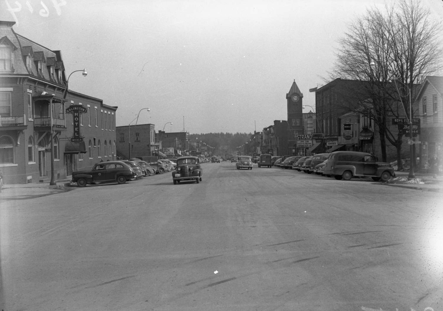 Black and white street view of Broadway Street in Tillsonburg. A sign for the Arlington Hotel is on the hotel to the far left. Store and business signs are hung on the front of buildings along the street. The post office and its clock tower are visible in the background. Many cars are parked along the street.