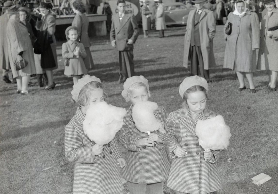 Three little girls wearing identical coats and hats eating cotton candy at a fair. A crowd of people are in the background. 