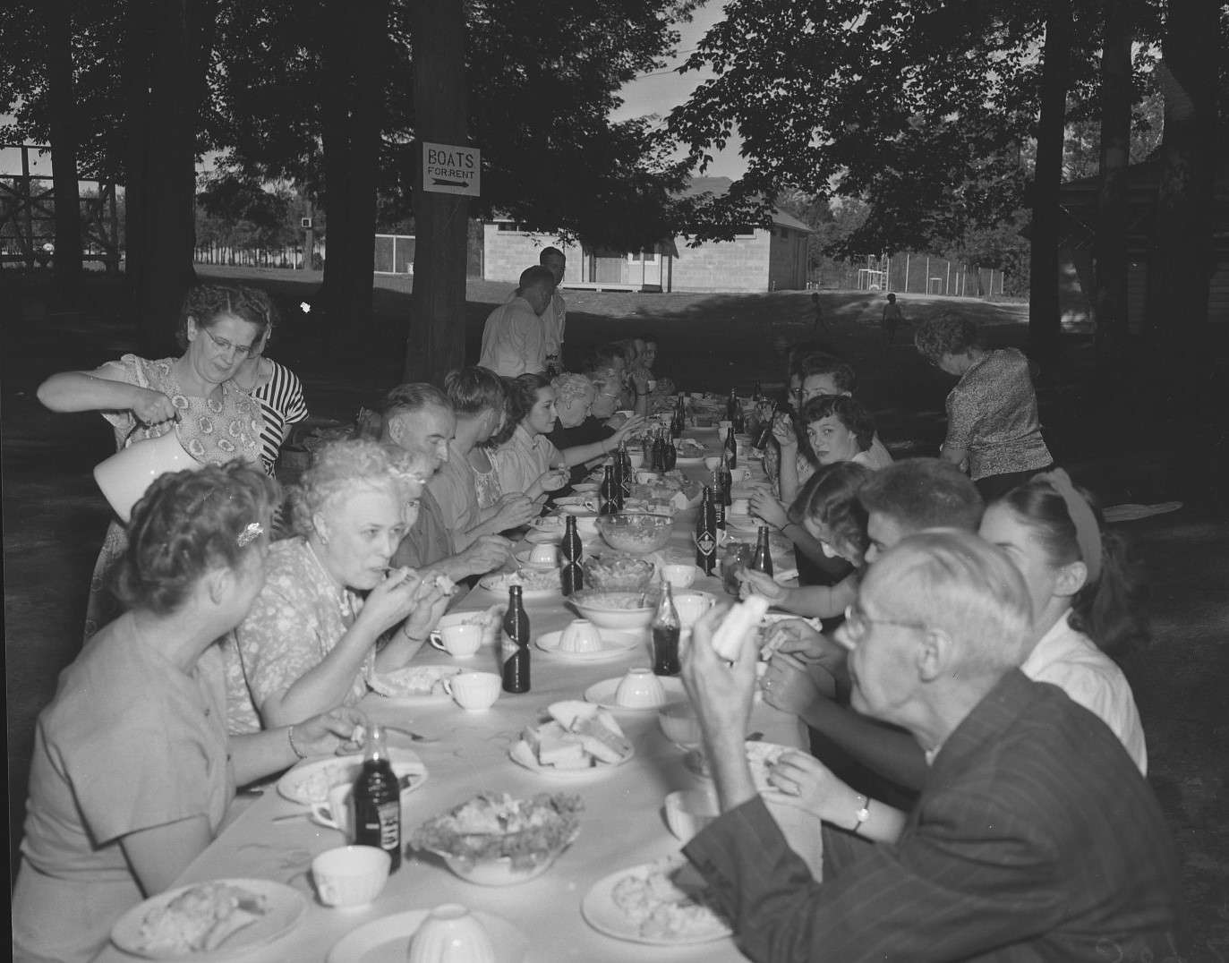 A large group of people sitting at a long table eating a picnic in a park filled with trees. A wooden building is in the background with a frost fence.