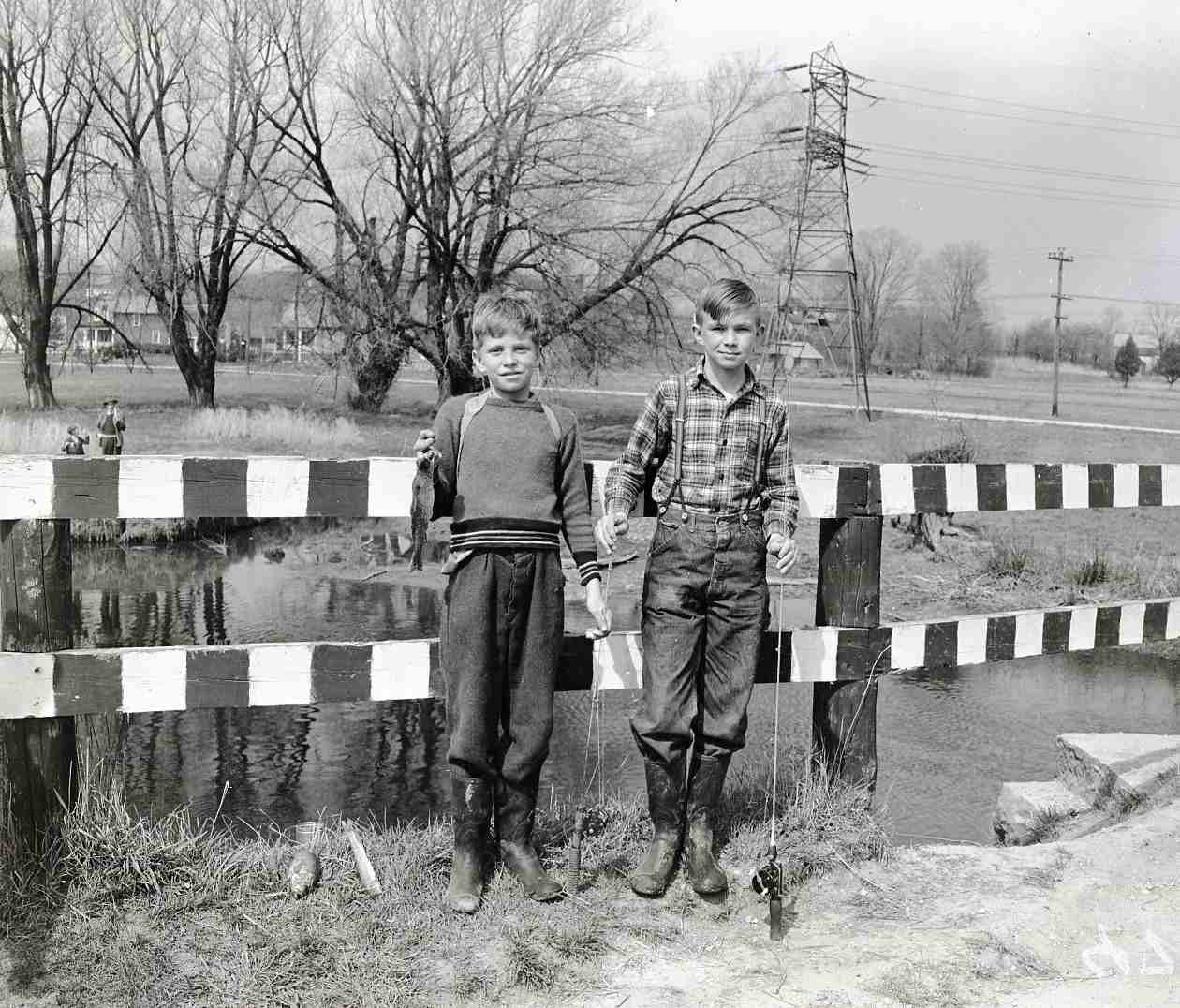 Two boys are holding fishing rods, with one of the boys holding a fish in his right hand. The boys are standing in front of a guard rail by a creek. Trees and hydro lines are in the background. Both of the boys are wearing rubber boots. The one boy is wear a sweat and trousers. The other boy is wearing jeans, with suspender and a plaid buttoned shirt.