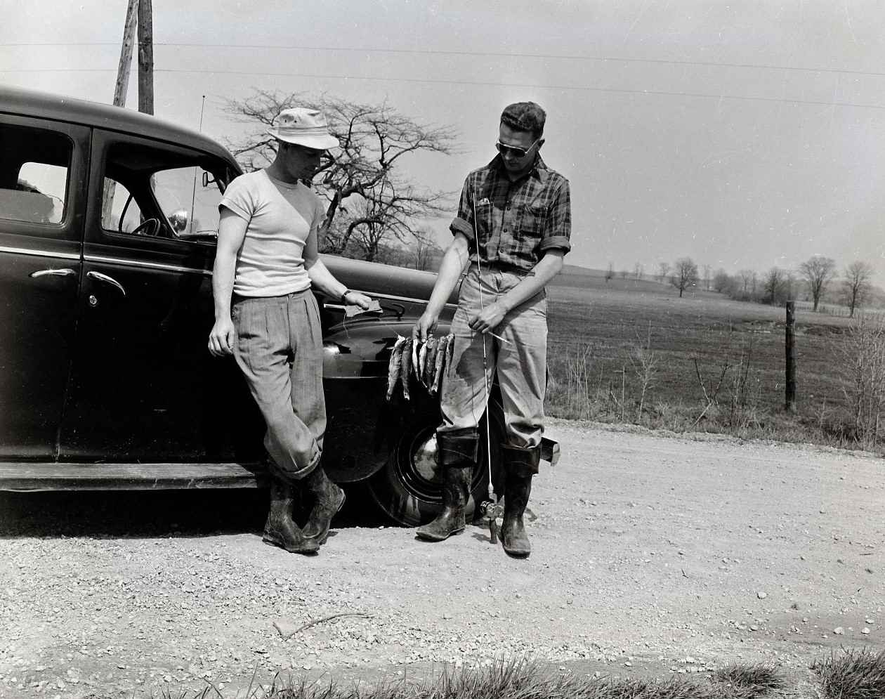 Two men posing in front of an automobile with a string of small fish they caught. They are both wearing rubber boots. The one man is wearing pants with a plaid buttoned shirt, sleeves rolled, up and sunglasses. The other man is wearing trousers with a light coloured t-shirt and hat.