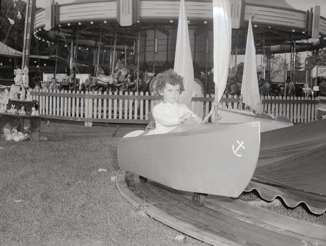 A young child is sitting in a boat ride at a fair, with a merry-go-round and strongman game in the background.