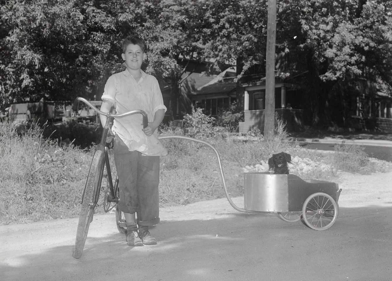 A boy is standing with a bicycle with a small wagon attached to the back of the bike. A dog is sitting in the wagon and has its tongue out.