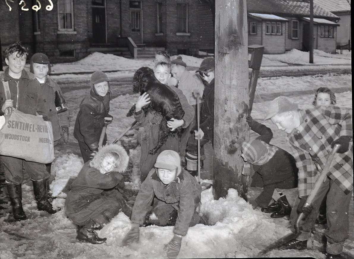 Group of children wearing winter clothing and playing in the snow on the side of the street around a hydro pole. Some of the children are using shovels in the snow. One boy is wearing a large bag around his torso that reads 