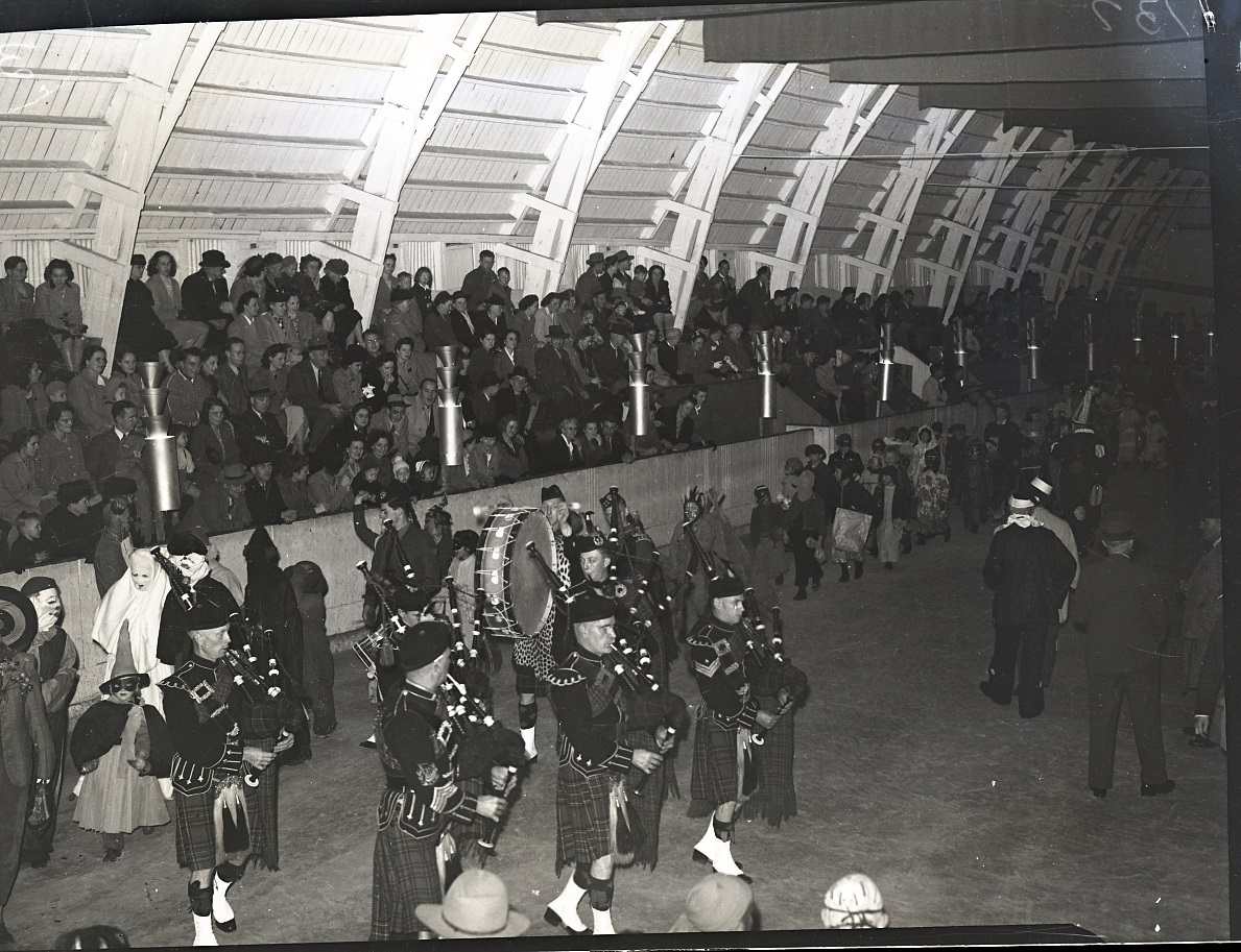 Photograph of the Ingersoll Pipe Band at the Ingersoll Community Centre, performing for the Halloween Frolic. A crowd of people are sitting in the stands, and children are wearing costumes in a parade. The pipers are wearing uniforms and carrying instruments including bag pipes and drums.