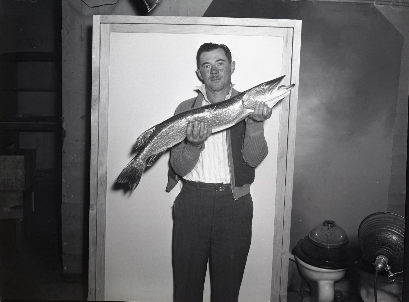 A man is posing in front of a white screen holding up a large pike fish he caught.