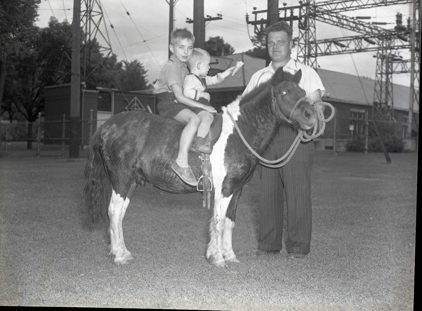 Two young boys are sitting on a pony. A man is holding the pony with w lead. The smallest boy is handing an ice cream cone to the man holding the pony.