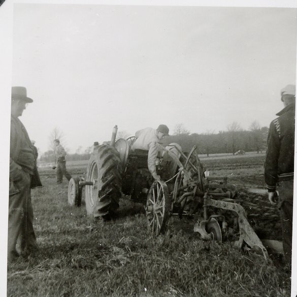 A young man sits on farm equipment in a field while older men around him watch.