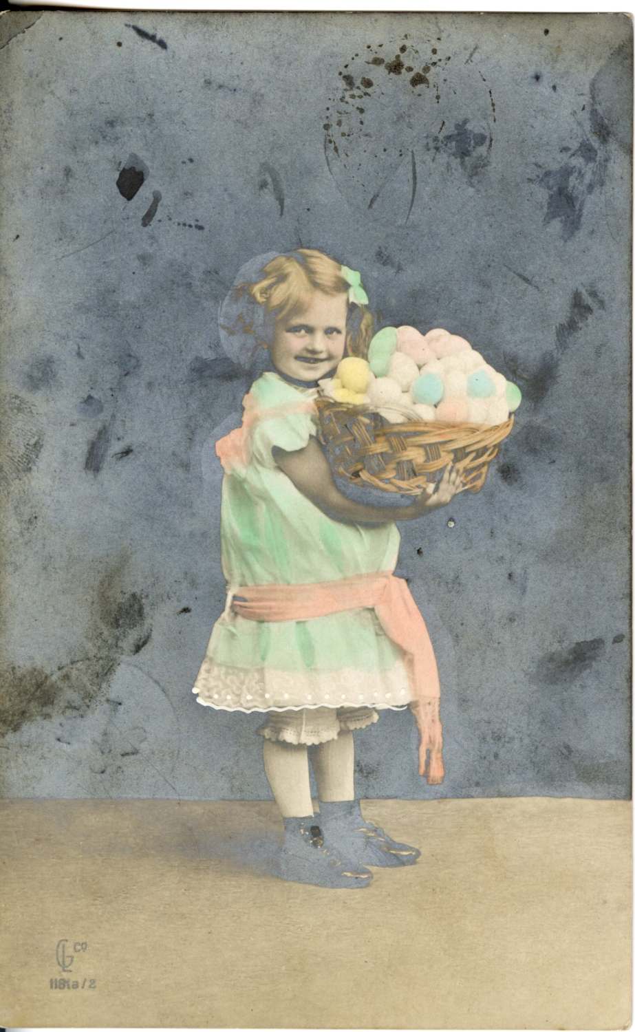 Postcard of a little girl holding a basket of Easter eggs. The image is colourized. The girl is wearing a mint green dress with a peach coloured ribbon.