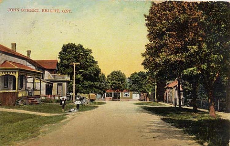 A colour postcard with a view of John Street in Bright, Ontario. The street is not paved, and has several homes and wooden hydro poles lining it. Children are standing on the side of the street.