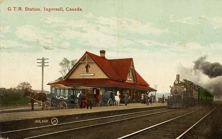 A postcard with a photograph of the Grand Trunk Railway Station in Ingersoll, which has been colourized. The station is tan in colour with a rust-coloured roof. Several horse-drawn buggies stand on the station platform along with people waiting for the train. A train is pulling up to the station.