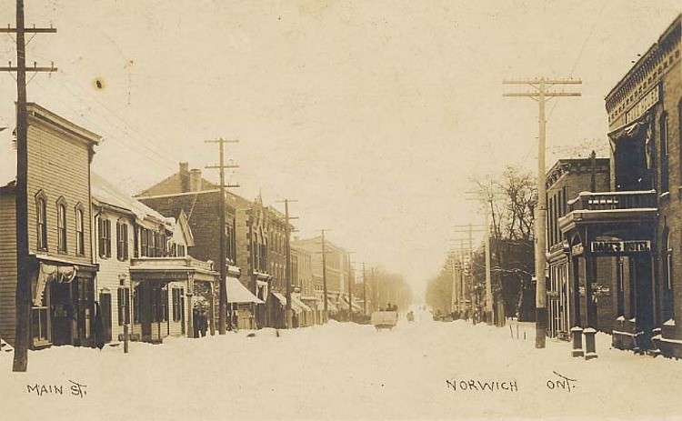 A photograph of the main street in Norwich, Ontario. Wooden frame and brick buildings line the street along the several telephone poles. A horse-drawn wagon is in the distance travelling down the street.