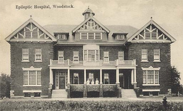 A postcard featuring a black and white photo of the former Epileptic Hospital in Woodstock, Ontario. The hospital is a large brick building. Three nurses are standing on the front verandah.