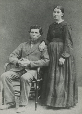 Portrait of John and Lydia Zurbrigg. John is sitting in a chair wearing a suit, Lydia is standing behind him with her hand on his shoulder, wearing a dark dress.