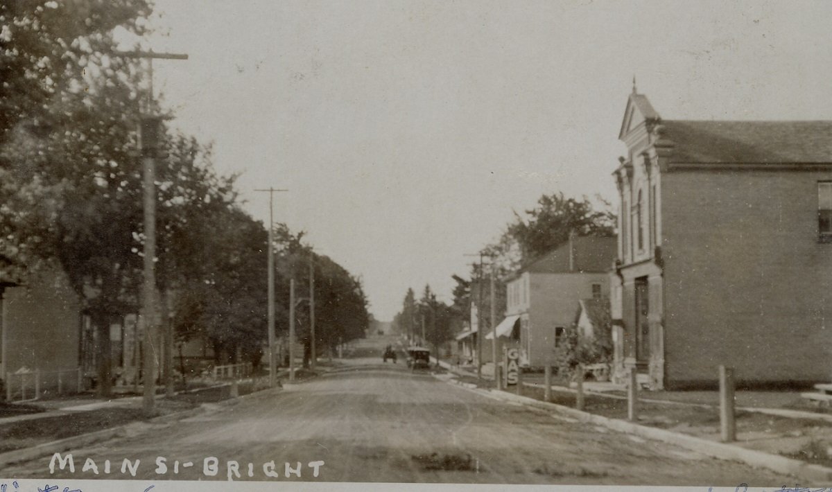 a black and white photo of Main Street in Bright, Ontario from 1925. The dirt street has several buildings lining it and light and hydro poles. On the right side is a gas station. Two automobiles can be seen in the distance.