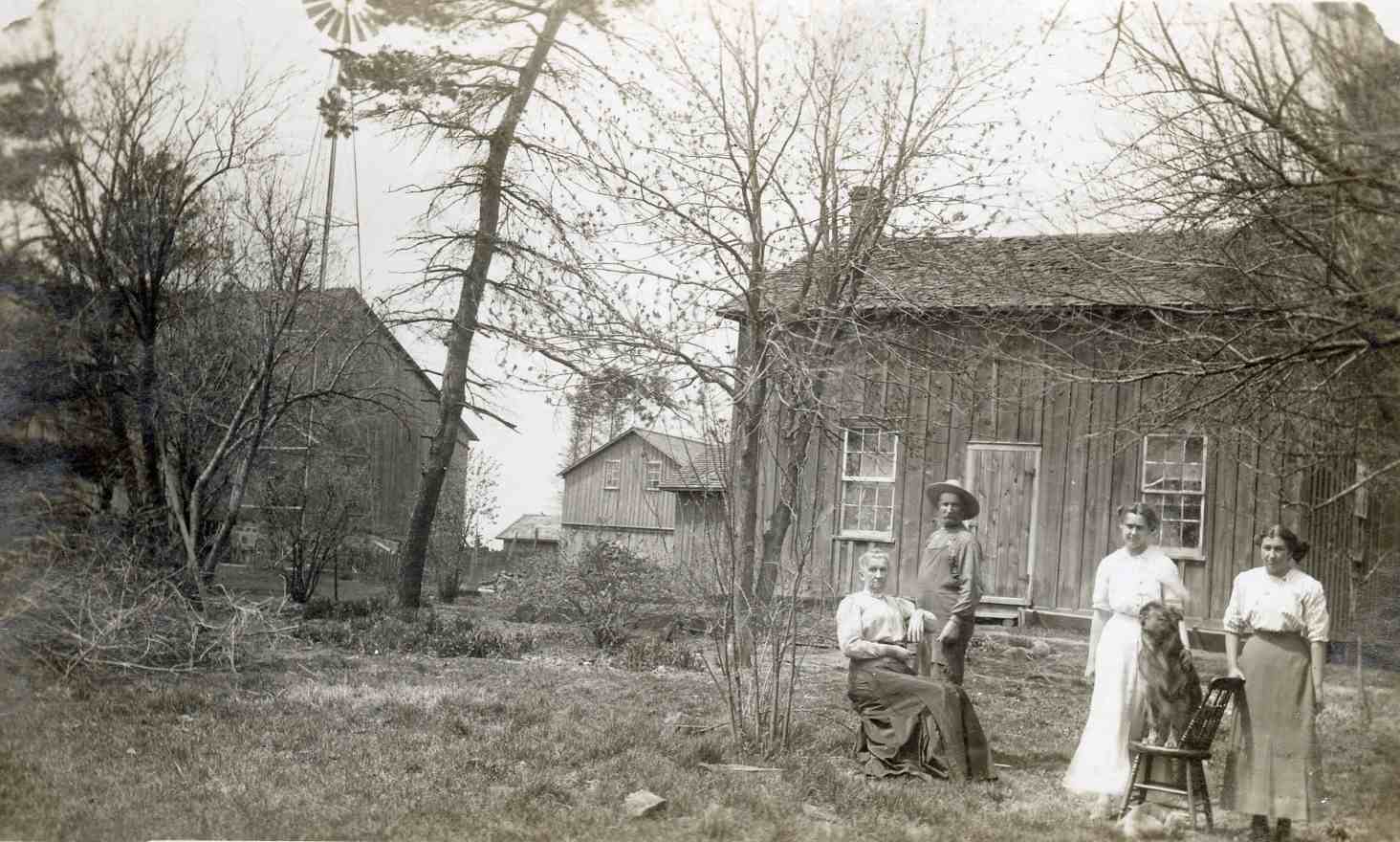 Four people are posing on a farm, one man and three women. The women are wearing white blouses and long skirts. One woman is seated in a chair. The man is wearing a hat and overalls. Wooden farm buildings and a windmill are in the background. A shaggy dog is seated on a chair with the group as well.