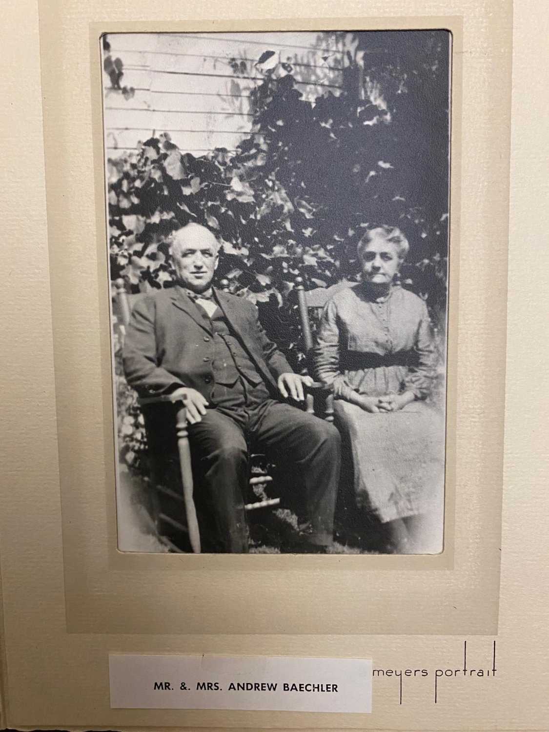 A photo of Mr. and Mrs. Andrew Baechler. They are more mature in age, and are sitting in wooden rocking chairs at the side of a house covered in foliage.