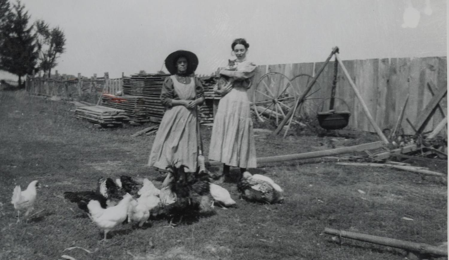 Two women are standing in a farmyard wearing long dresses and aprons. One woman has a large-brimmed hat on her head. The second woman is holding a cat in her arms. Behind the women are piles of planks of wood. There is also a large pot hung over a fire. A group of chickens are standing at the women's feet. The women are making soap in the pot.