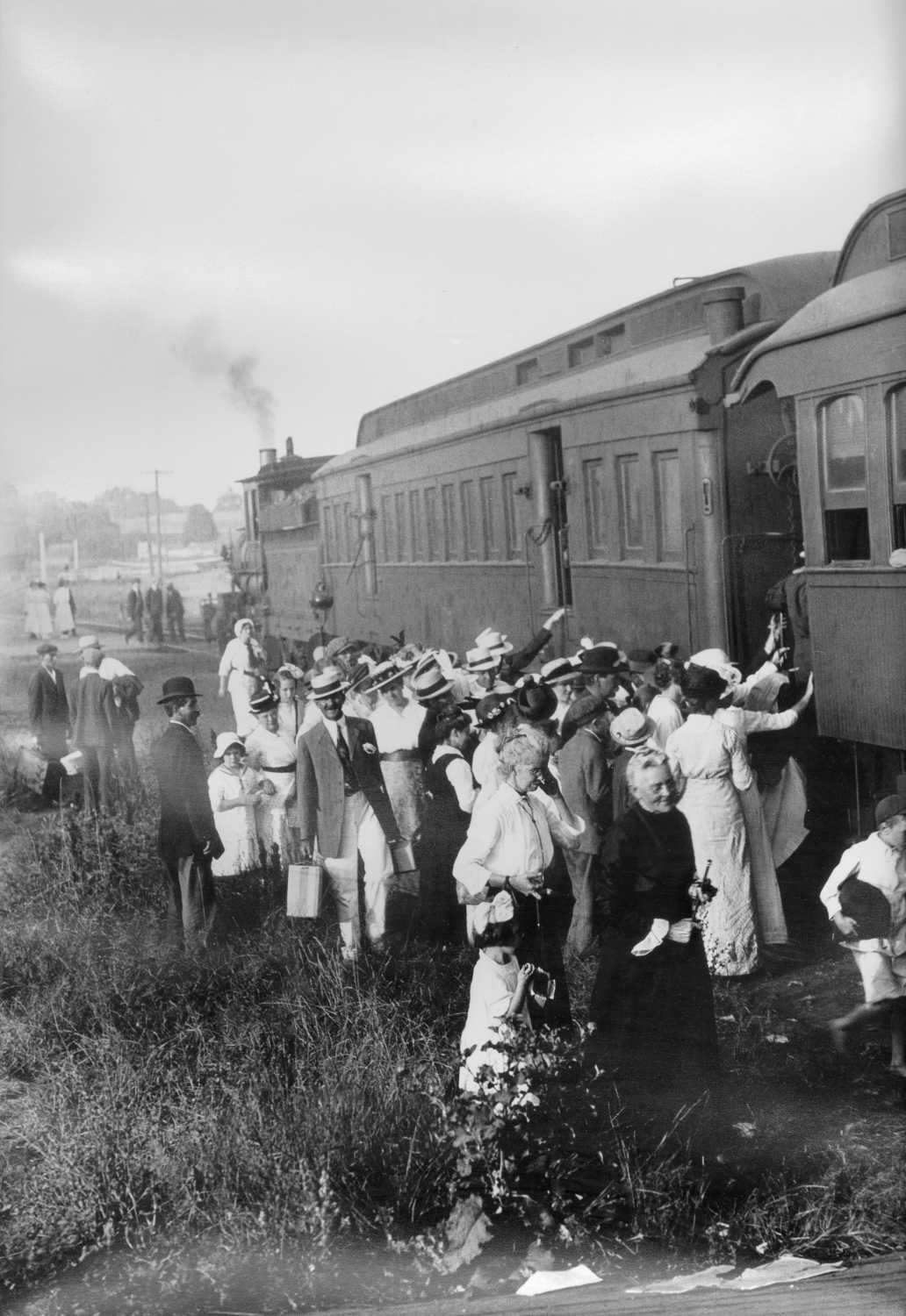 People gather in a large group beside a train, waiting to board.