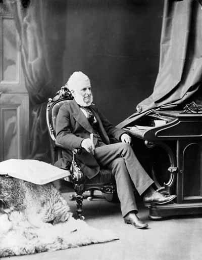 Sir Francis Hincks sitting in a chair at a desk for a studio portrait.