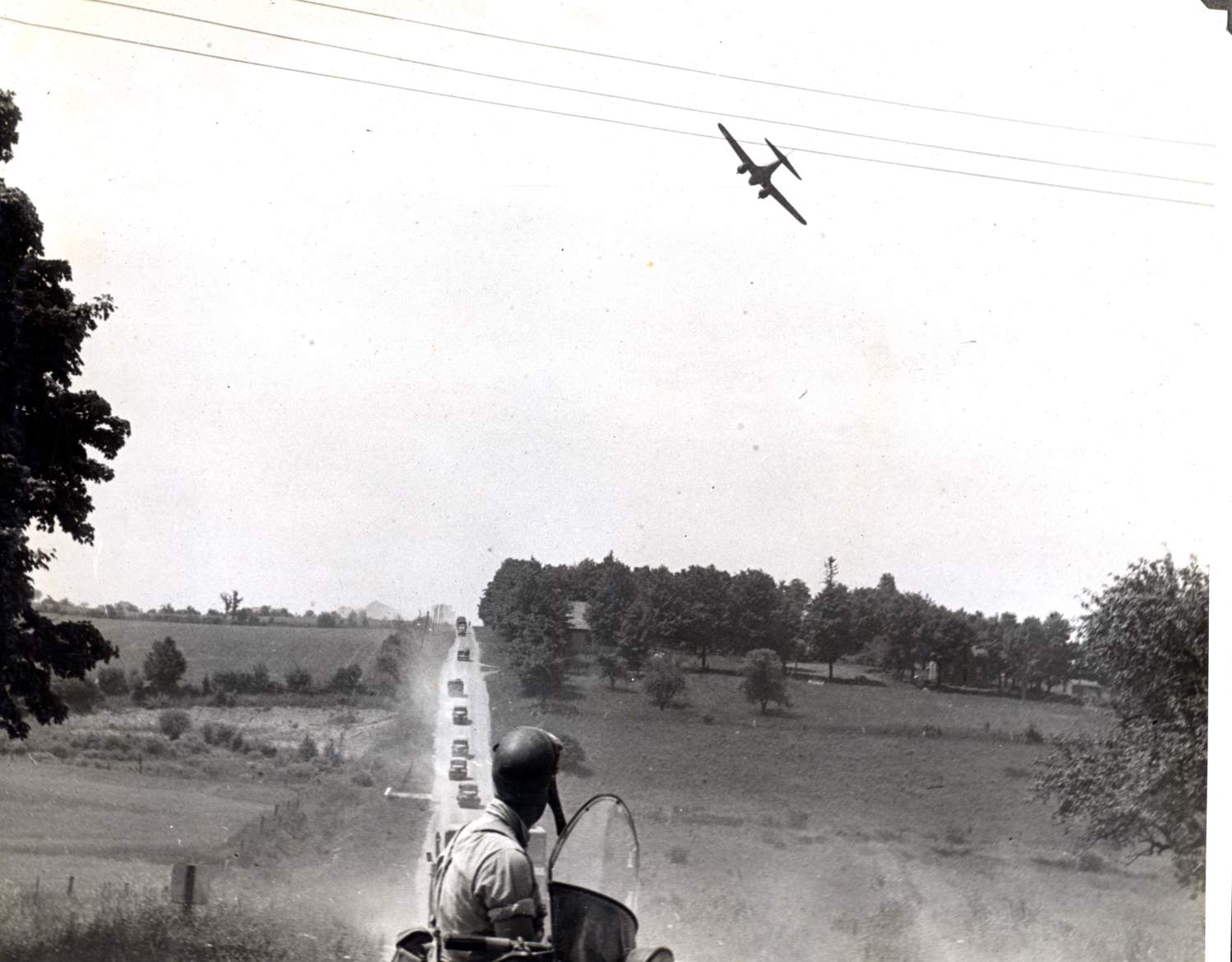 A man sitting on a motorcycle looking up at a small plane in the sky. A long line of military vehicles are driving along the dirt road ahead of him.