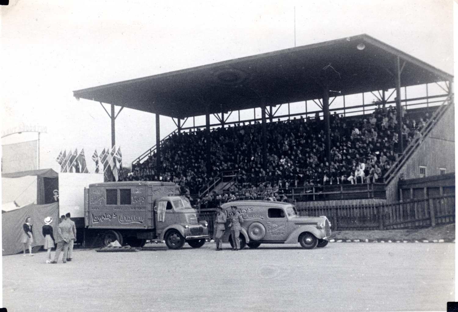 A large crowd of people are sitting in the grandstands at the Woodstock Driving and Maintenance School. On the ground in front of the stands there is a truck and automobile parked that have 