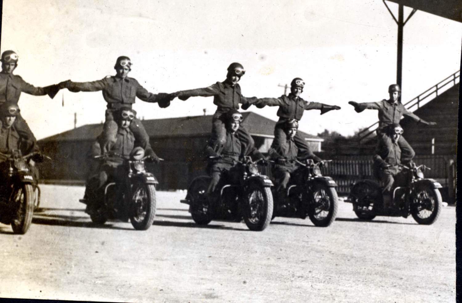 Five men in uniform ride motorcycles alongside each other at the Woodstock Driving and Maintenance School. Each driver has a man sitting on his shoulders, holding hands with the man beside him. They are all wearing helmets with goggles.