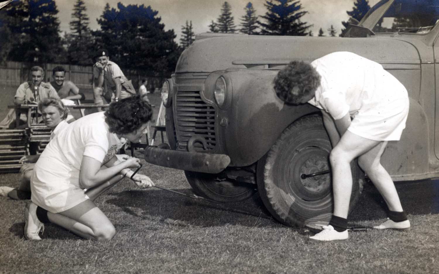 Two women with tire irons change a tire on a military vehicle at the Woodstock Driving and Maintenance School. Men in uniform are sitting in the background observing.